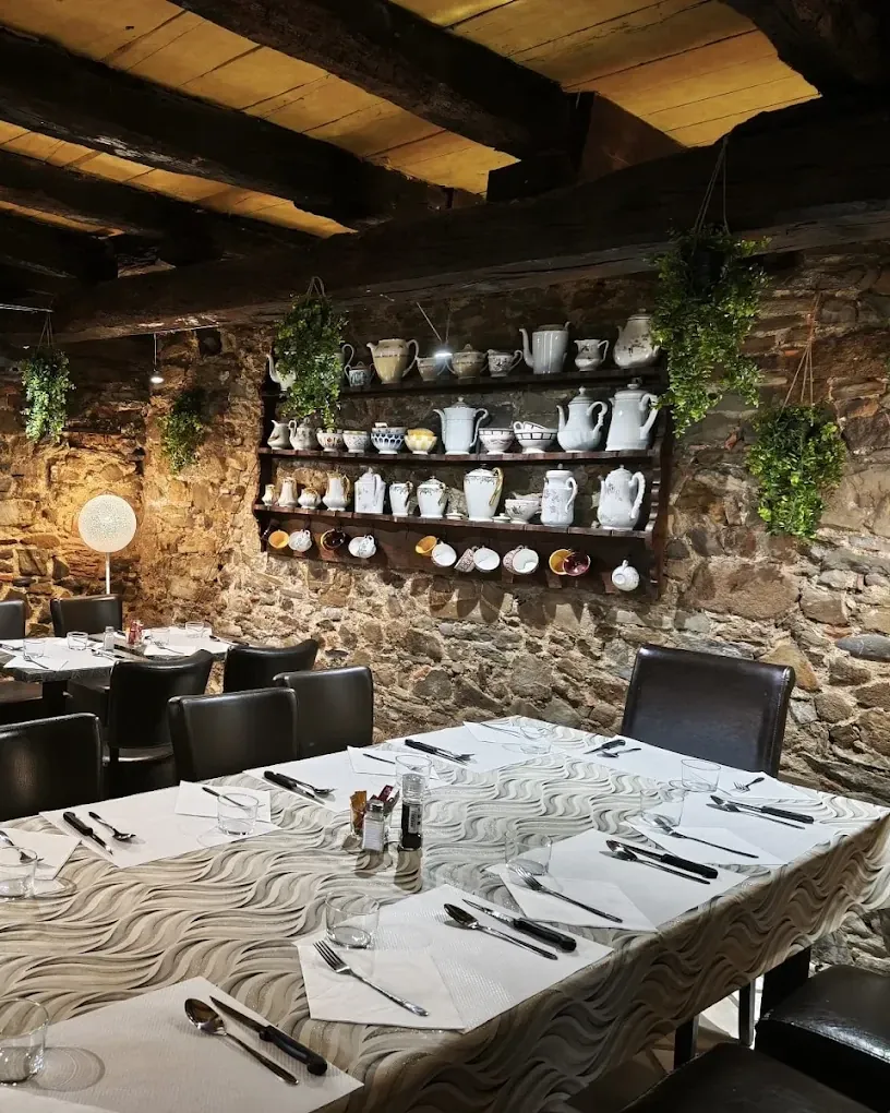 La salle de La Table de Bastida : murs en pierre, poutres apparentes, étagère de théières et de pichets anciens au-dessus d’une grande tablée dressée.